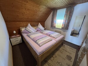 A cozy bedroom with a wooden ceiling, a bed with striped bedding, a nightstand, and a window with curtains.