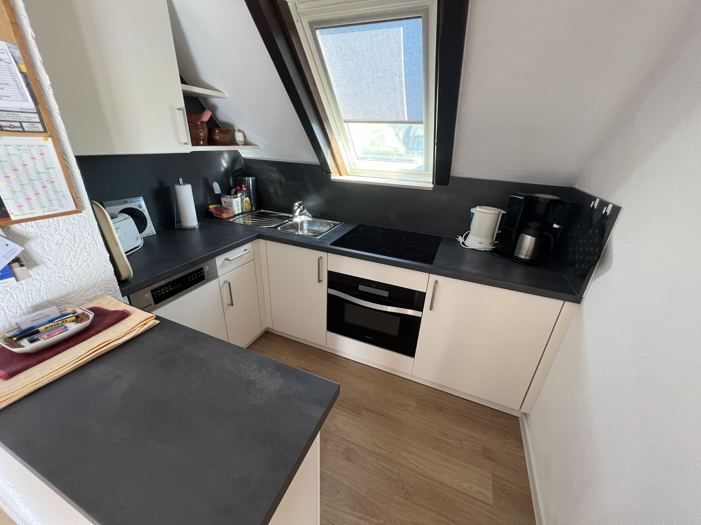 Een moderne keuken met een zwart aanrecht, witte kasten, een ingebouwde oven, een koffiezetapparaat en een raam boven de spoelbak.