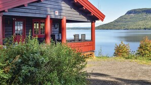 A cabin with a red roof and a wooden exterior, situated by a lake with a view of a forested hill in the background. - Amazing home in Vikersund with sauna (Vikersund)