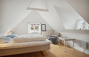 Quarto com teto inclinado, cama com roupa de cama branca, cômoda, cadeira de vime, mesa pequena e janela com vista para o exterior.