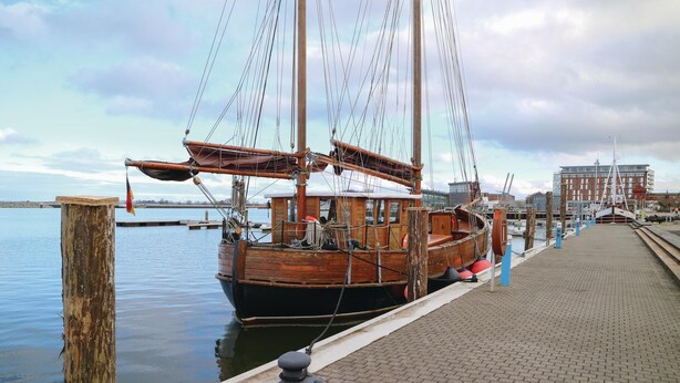 A wooden sailing boat docked at a pier with a cityscape in the background.