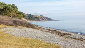 Un paisaje costero con una playa de guijarros, zonas verdes y una colina distante con una edificación.