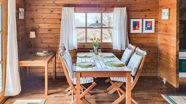 Interior de una cabaña de madera con una mesa de comedor preparada para cuatro comensales, una ventana con cortinas y obras de arte enmarcadas en las paredes.