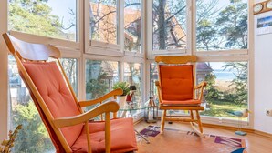 Et værelse med store vinduer, to trægyngestole med orange puder, et glasbord med en plante, og udsigt til det fri.