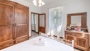 A bedroom with a large wooden wardrobe, a bed with white bedding, a bedside table with a mirror, and a window with sheer curtains.