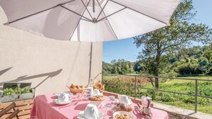 An outdoor dining area with a table set for breakfast, including bread, pastries, and a coffee machine, overlooking a scenic landscape.