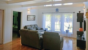 Uma sala de estar com sofá, mesa de centro e aparador para TV. Há uma janela com cortinas e um quadro na parede.