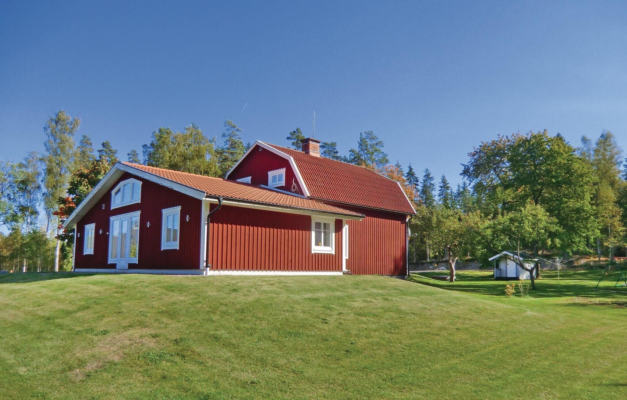 A red wooden house with a sloped roof, set on a grassy hill with trees in the background.