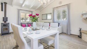 A dining area with a white table set for four, a grey sofa with pink cushions, a vase with flowers, and a large window with curtains. - Lovely home in Cubani (Cubani)
