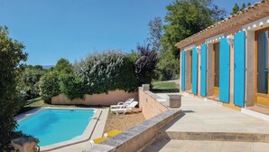 Una piscina con sillas de descanso, un porche cubierto con contraventanas azules y vistas al follaje circundante.