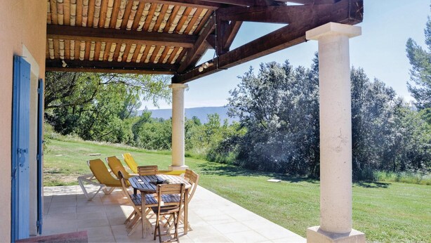 Un patio al aire libre con una pérgola, una mesa con sillas y vistas a un área verde con árboles y montañas al fondo.