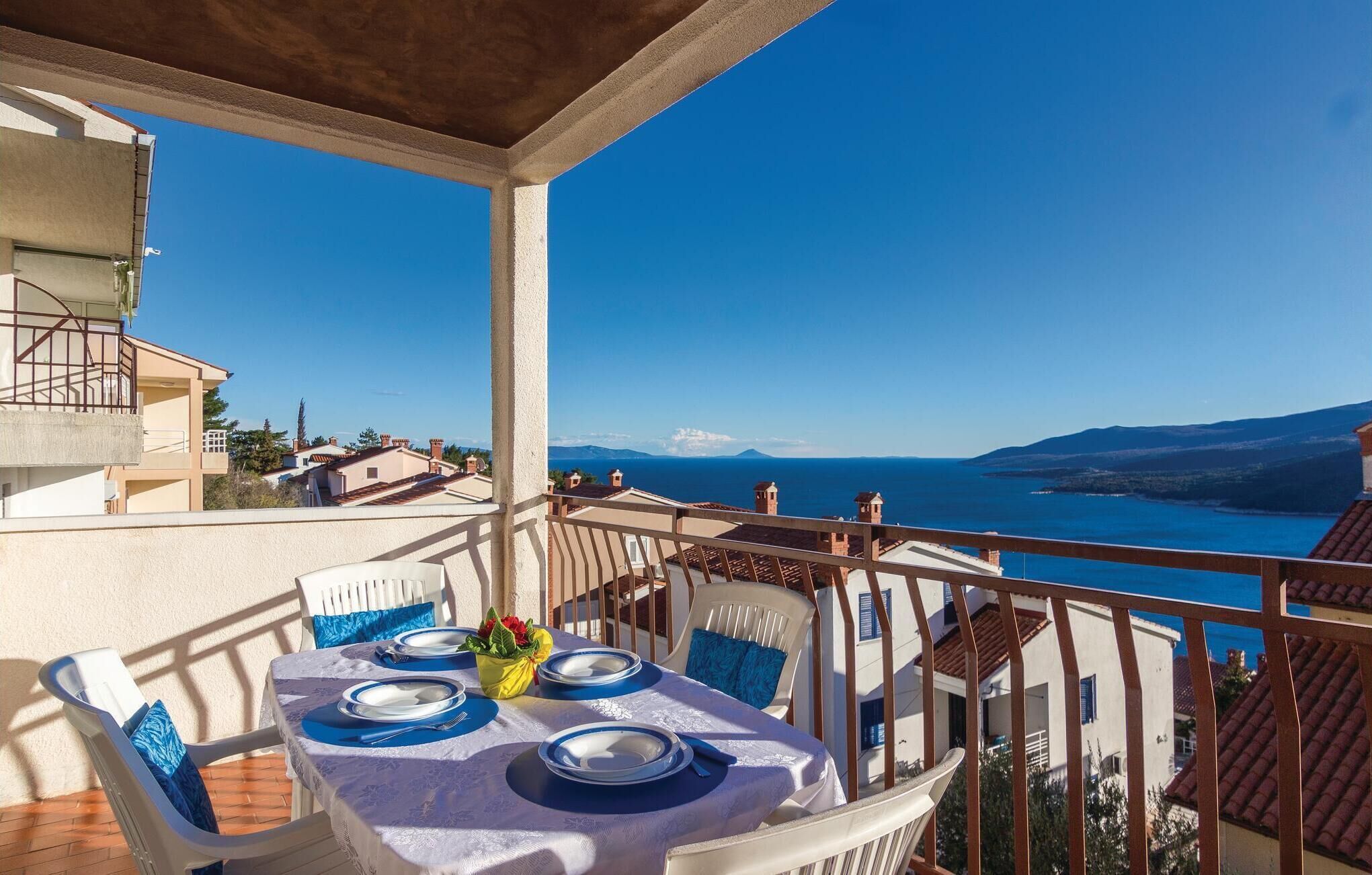 A balcony with a table set for dining, overlooking a coastal view with houses and mountains in the distance.