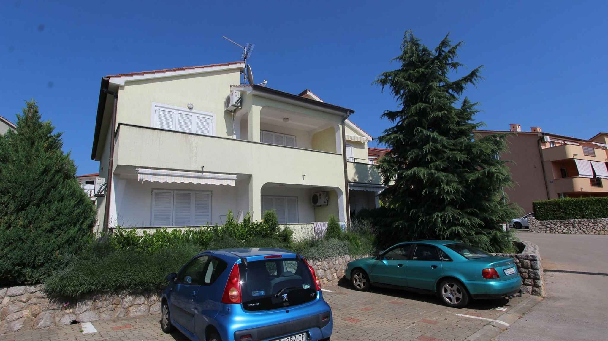 A two-story residential building with a balcony, parked cars, and a large evergreen tree.