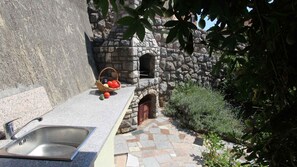 Un four en pierre, un évier et un panier de fruits sur une terrasse carrelée.