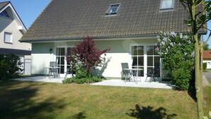 A two-story house with a tiled roof, a small garden with a tree and shrubs, and a patio with outdoor furniture. - Apartment Wiesenblick 1-1 bedroom up to max. 2 pers. - Vacation home Wiesenblick (Karlshagen)