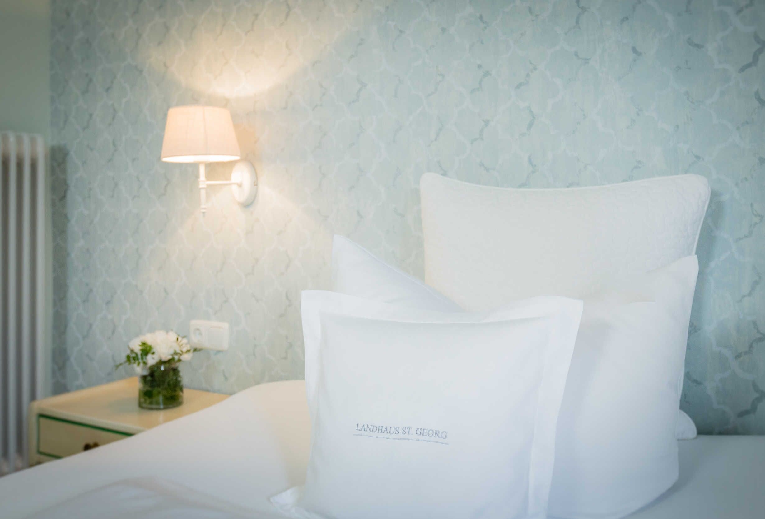 Ein ordentlich bezogenes Bett mit weißen Leinen und Kissen, eine Nachttischlampe und ein kleiner Blumenstrauß auf einem Nachttisch.