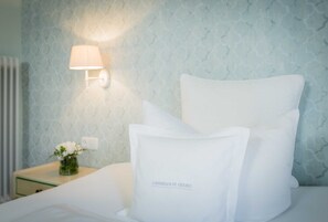 Una cama bien hecha con sábanas y almohadas blancas, una lámpara de noche y un pequeño florero con flores sobre una mesita.