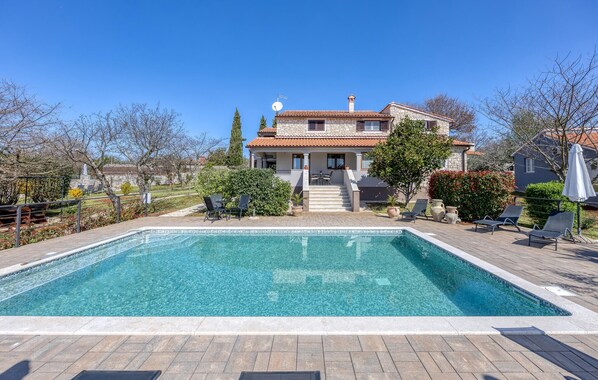 Un área de piscina con una piscina azul cristalina, una casa de piedra con techo de tejas y una escalera que conduce a la casa.