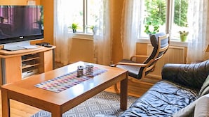 Een woonkamer met een televisie, een houten salontafel, een kamerbreed tapijt met dessin, een zwarte leren bank en een raam met gordijnen.
