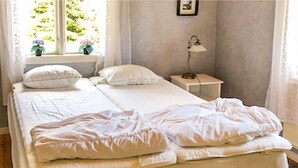 Een net opgemaakt bed met witte lakens en kussens, een nachtkastje met een lamp, en een raam met gordijnen en een klein plantje.