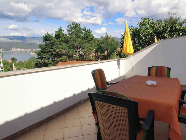 Une terrasse extérieure avec une table et des chaises, un parasol jaune, et une vue sur une étendue d’eau et des montagnes lointaines.