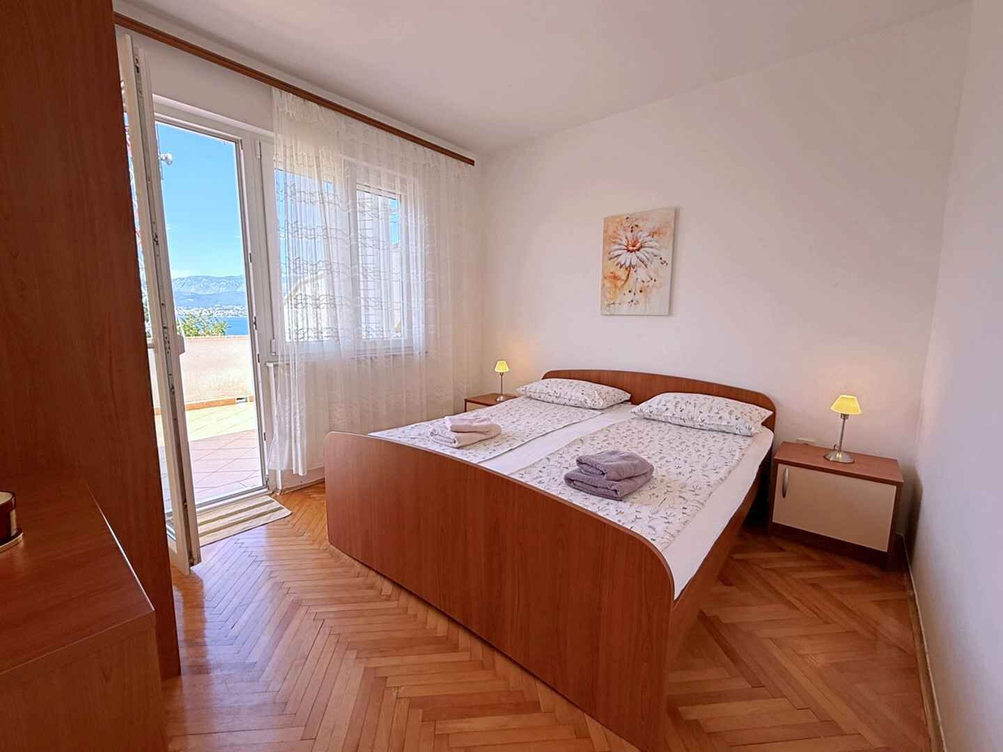 A bedroom with a wooden bed, two bedside lamps, a window with curtains, and a painting on the wall.