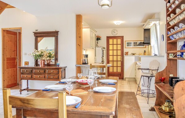 Comedor con mesa de madera dispuesta para cuatro, cocina al fondo y estante con vajilla.