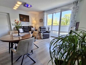 Ein modernes Wohnzimmer mit Essbereich, einer Sofagarnitur, einem Sessel, einem Tisch und einem großen Fenster mit Blick.