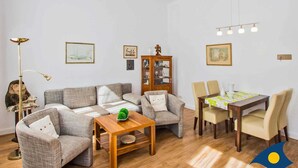 Ein Wohnzimmer mit einer Sofagarnitur, einem Holztisch, einem Esstisch mit Stühlen, einem Schrank und einer Stehlampe.