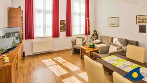 Ein helles Wohnzimmer mit einer Couch, einem Couchtisch und einem Esstisch. Große Fenster mit roten Vorhängen, eine Heizung und eine Stehlampe sind ebenfalls vorhanden.