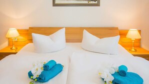 Ein Doppelbett mit weißen Leinentüchern, zwei weiße Kissen und zwei blaue Handtücher mit weißen Blumen. Auf hölzernen Nachttischen stehen jeweils zwei Nachttischlampen.