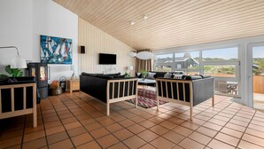 Ein modernes Wohnzimmer mit Kamin, schwarzen Sofas, einem Fernseher und einem großen Fenster mit Blick auf ein Haus.