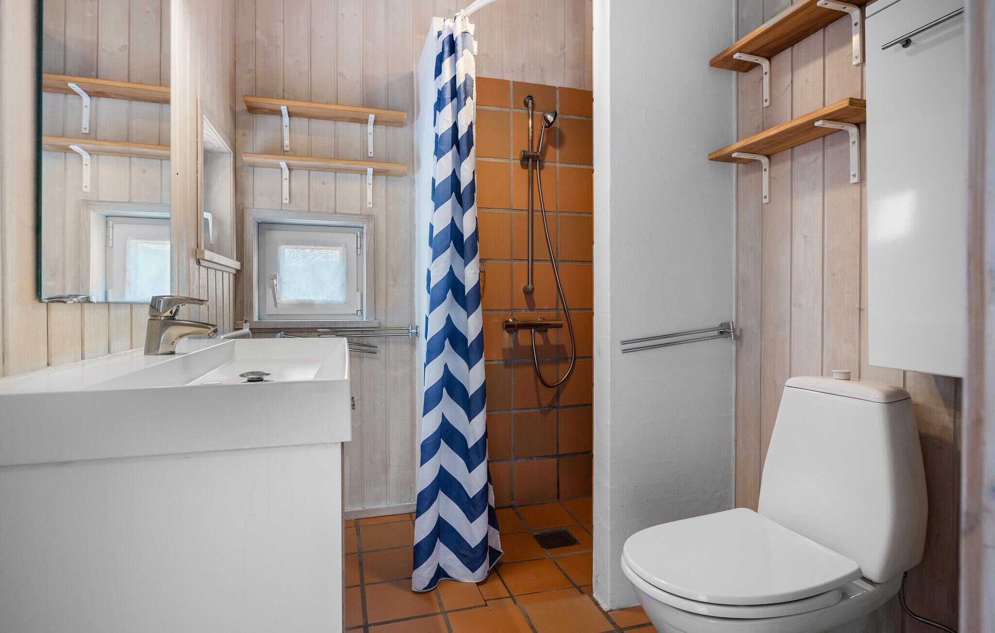 Un bagno con un lavandino bianco, un water, una doccia con tenda blu e bianca, ed alcune mensole in legno sopra il lavandino.