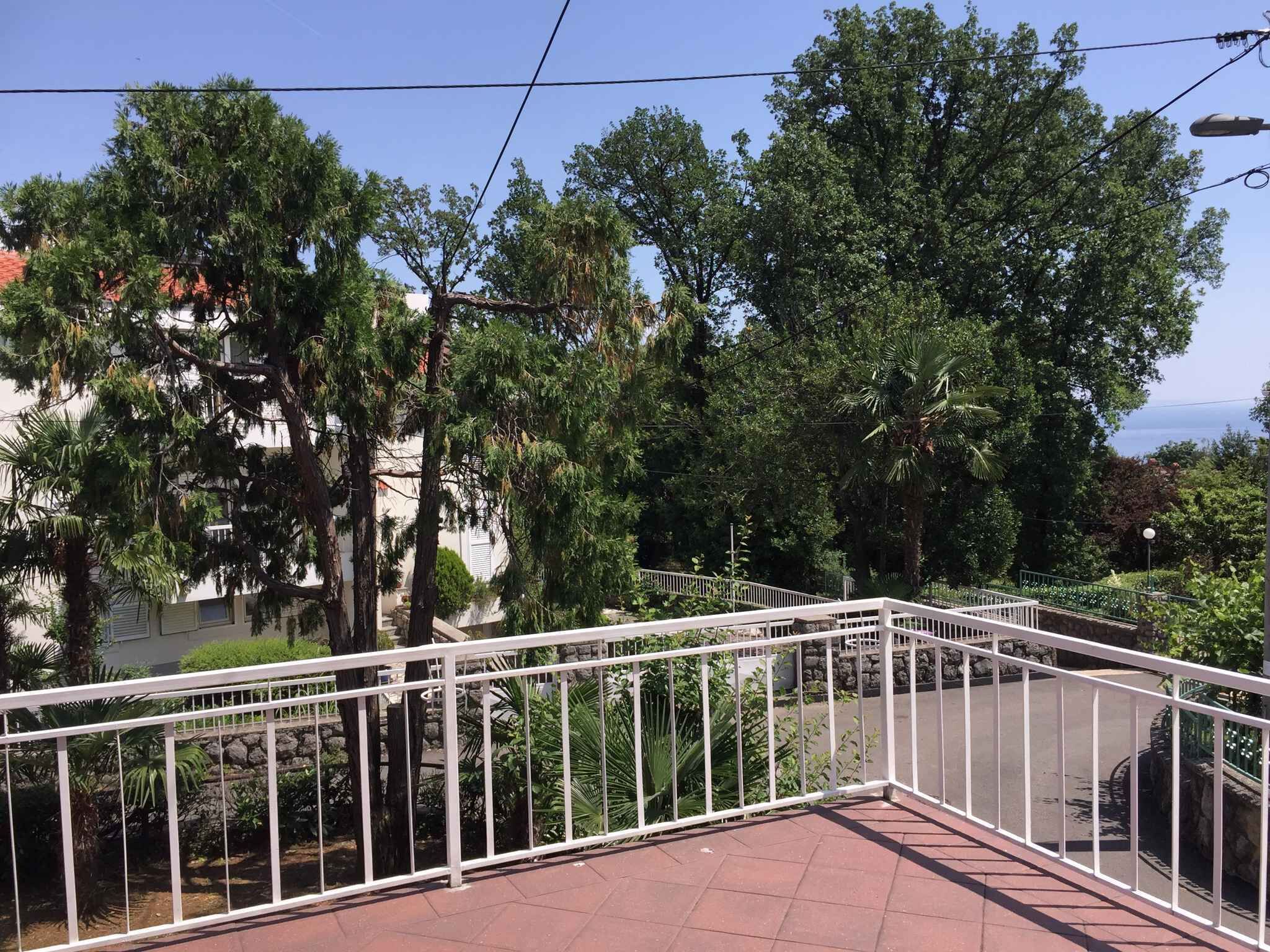 A balcony with a white metal railing overlooking a garden with various trees and a view of a building in the background.