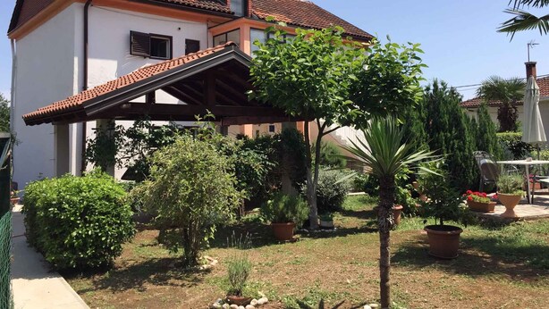 Una casa de dos pisos con techo de tejas, un porche cubierto y un jardín con diversas plantas y una palmera.