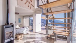 Una habitación de hotel moderna con cama, banca de madera, chimenea y un amplio ventanal con vistas a la montaña.