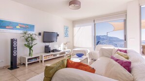 Una sala de estar moderna con un televisor de pantalla plana, un sistema de sonido, un sofá blanco y un amplio ventanal con vistas a la montaña.