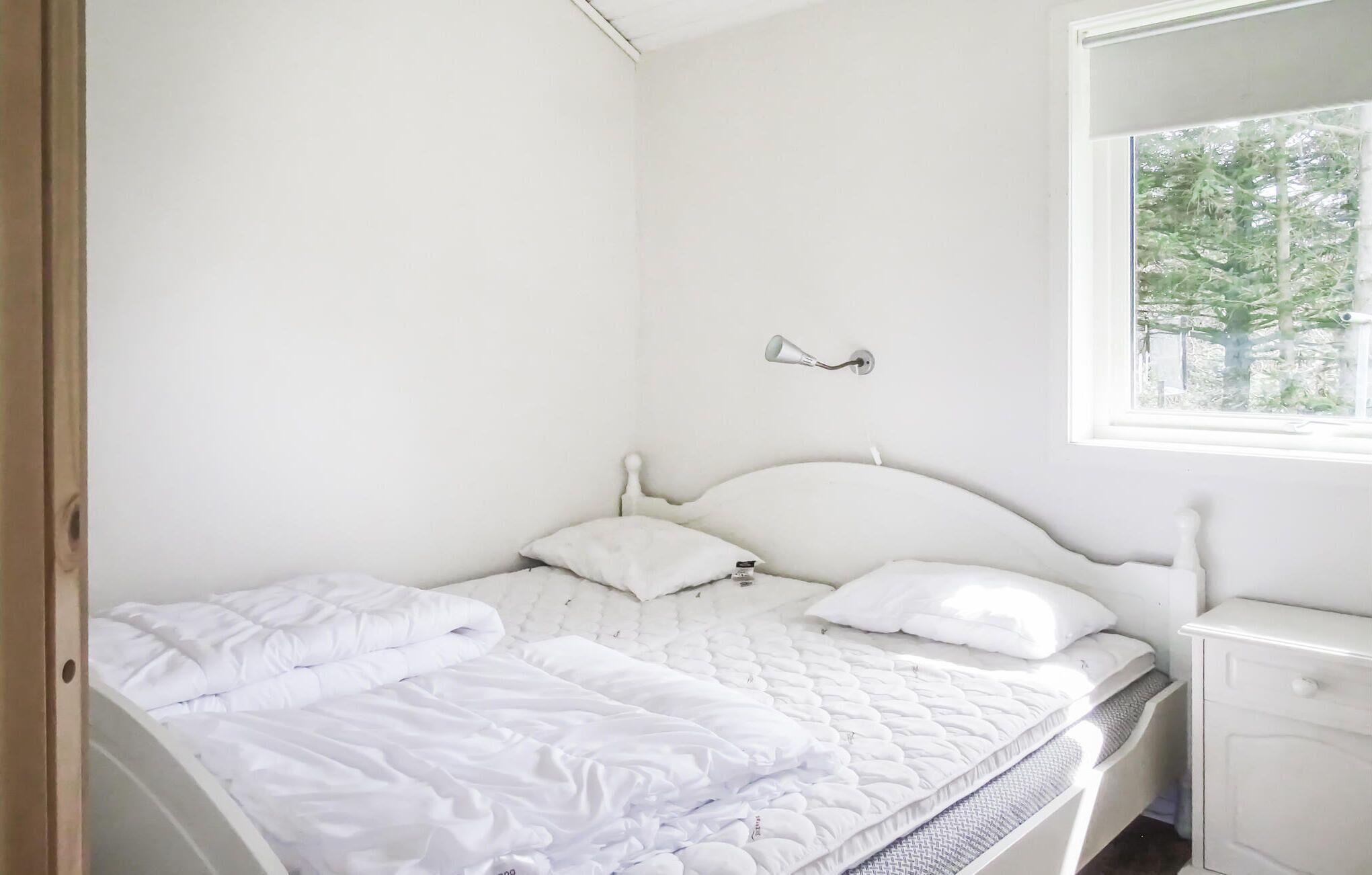 A white bedroom with a bed, a nightstand, and a window with a view of trees.