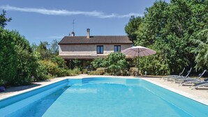 Ein rechteckiger Swimmingpool mit einer gefliesten Terrasse, umgeben von üppiger Bepflanzung und einem Haus mit einem geflasten Dach im Hintergrund.