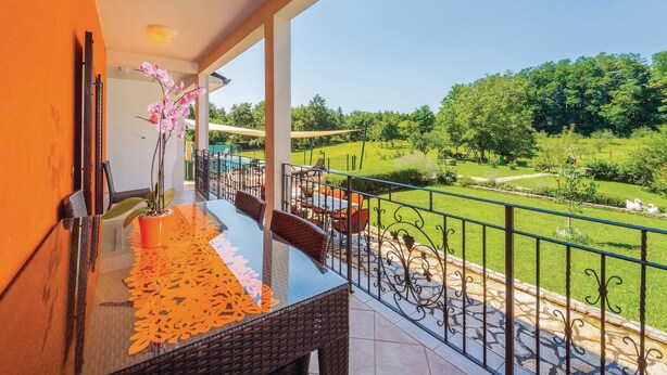 A balcony with a dining table, chairs, and a potted orchid. The balcony overlooks a green landscape with trees and a swimming pool. - Nice home in Nedescina (Nedescina)