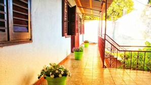A corridor with tiled flooring, potted plants, a staircase with a metal railing, and wooden shutters on the windows.