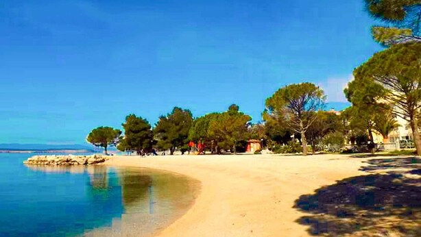 A beach with clear water, trees, and buildings in the background.