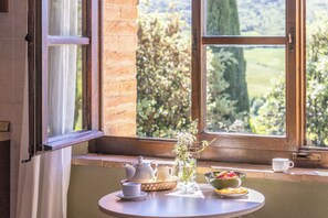 Una mesa con juego de té, un cuenco de fruta y un florero con flores junto a una ventana con vistas al verdor.