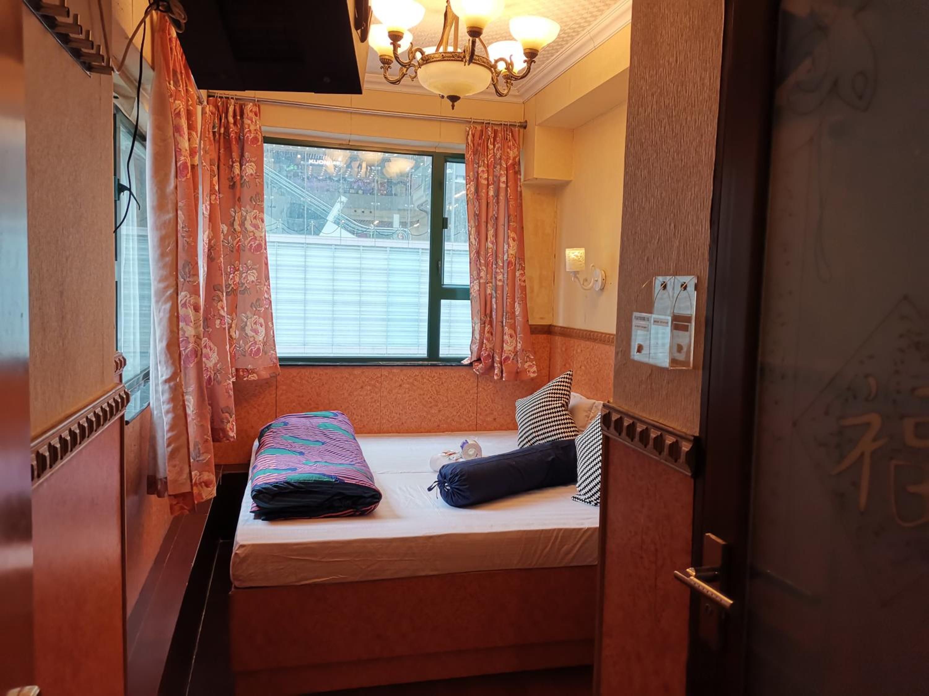 Een kleine, knusse kamer met een enkel bed, een raam met gordijnen, een kroonluchter en een deur met een bordje.