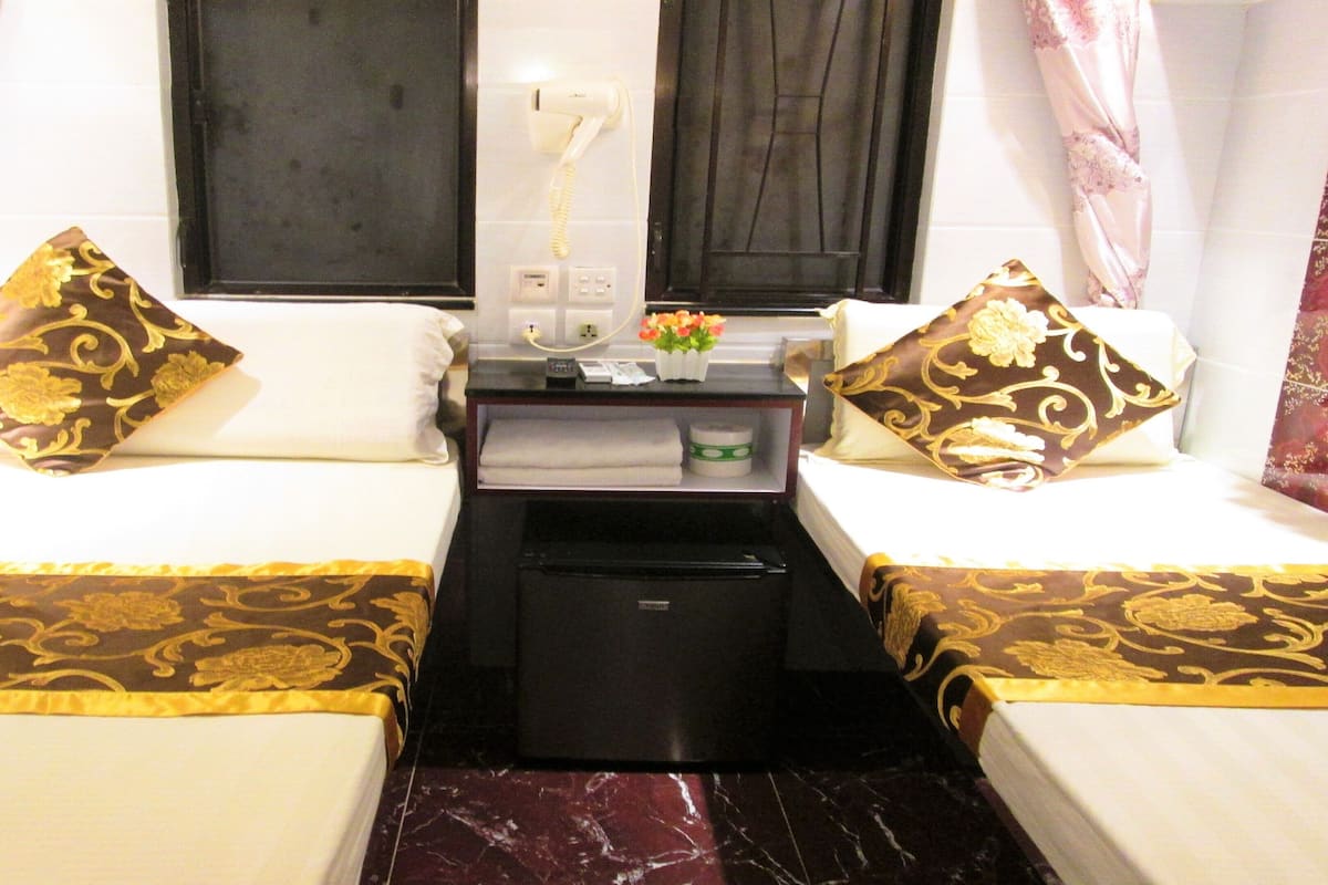 A hotel room with two beds, each with a patterned bedspread and matching pillows. There is a small black minibar and a hairdryer on a table between the beds.
