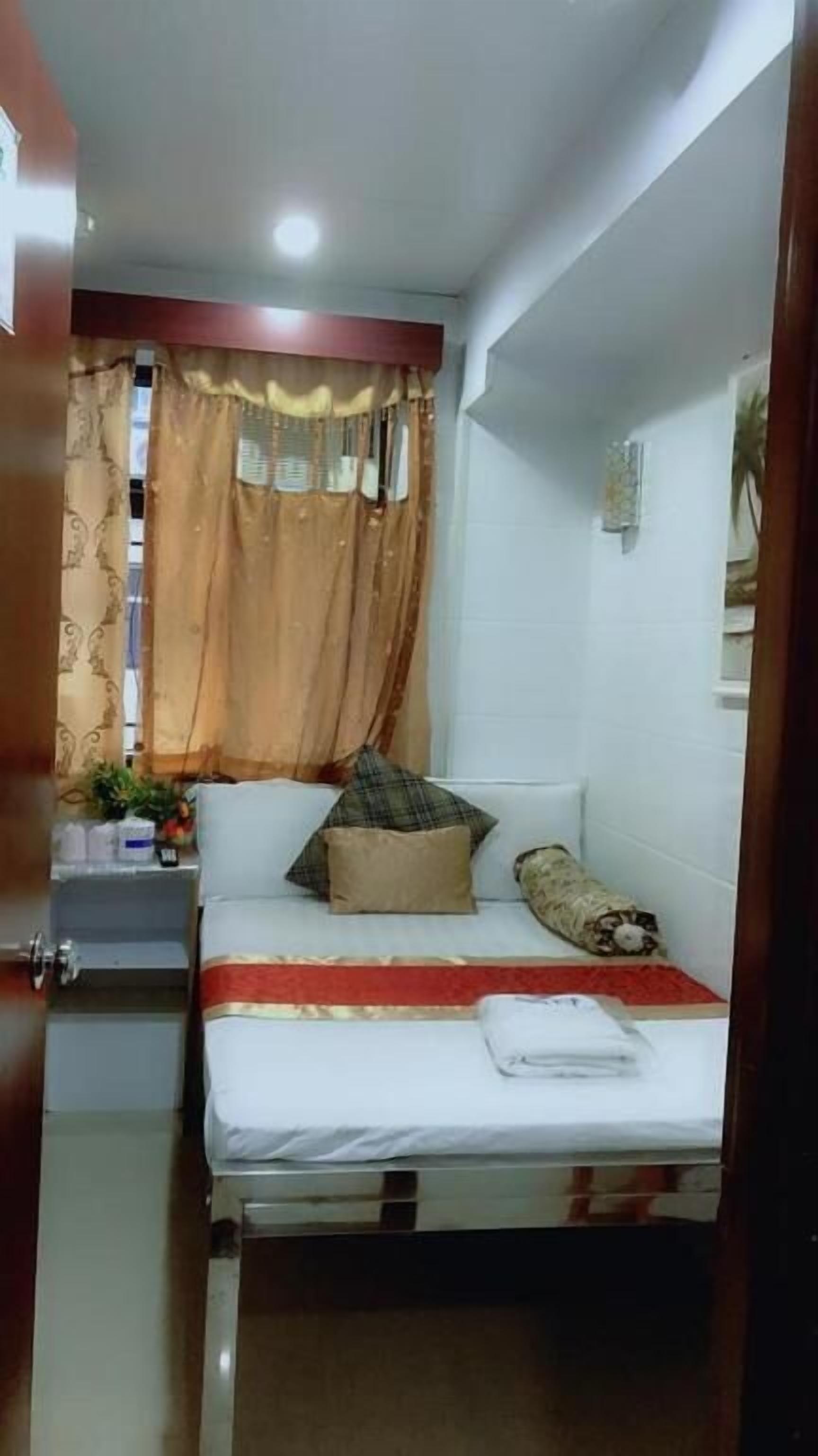 A small, compact room with a bed, a nightstand, a window with curtains, and a wall-mounted air conditioning unit.