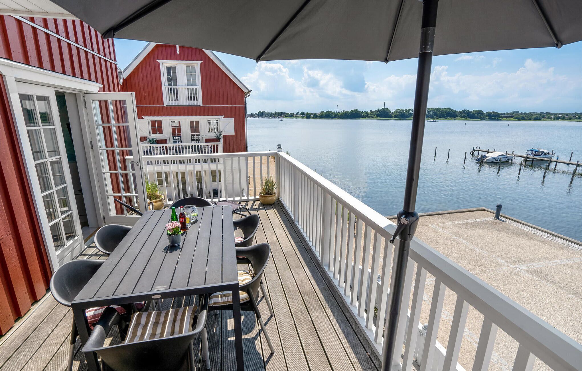 Une terrasse en bois avec une table et des chaises, donnant sur un lac paisible où l’on peut apercevoir des bateaux.