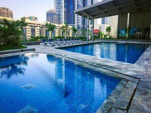 A swimming pool with lounge chairs, surrounded by palm trees and modern buildings in the background.