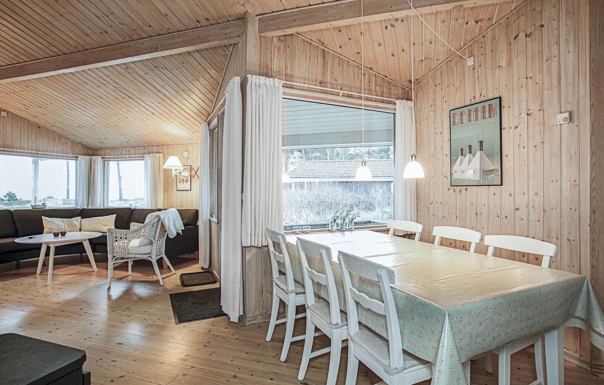 A cozy cabin interior with a wooden ceiling, a dining area with a table and chairs, a sofa, and a large window offering a view of the outdoors.
