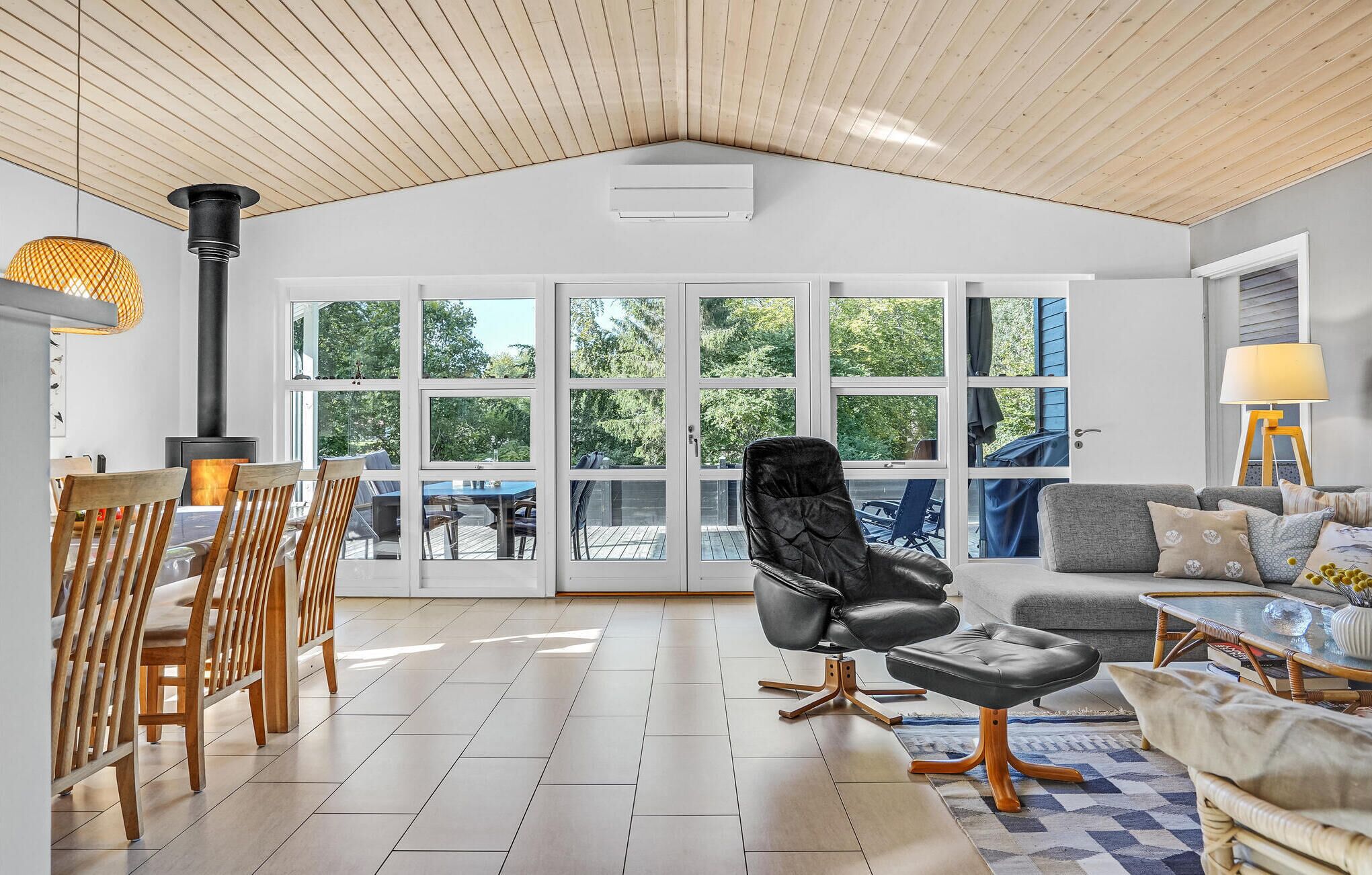 A spacious room with a high ceiling, large windows, a dining area with wooden chairs, a black leather chair, and a sofa with cushions.
